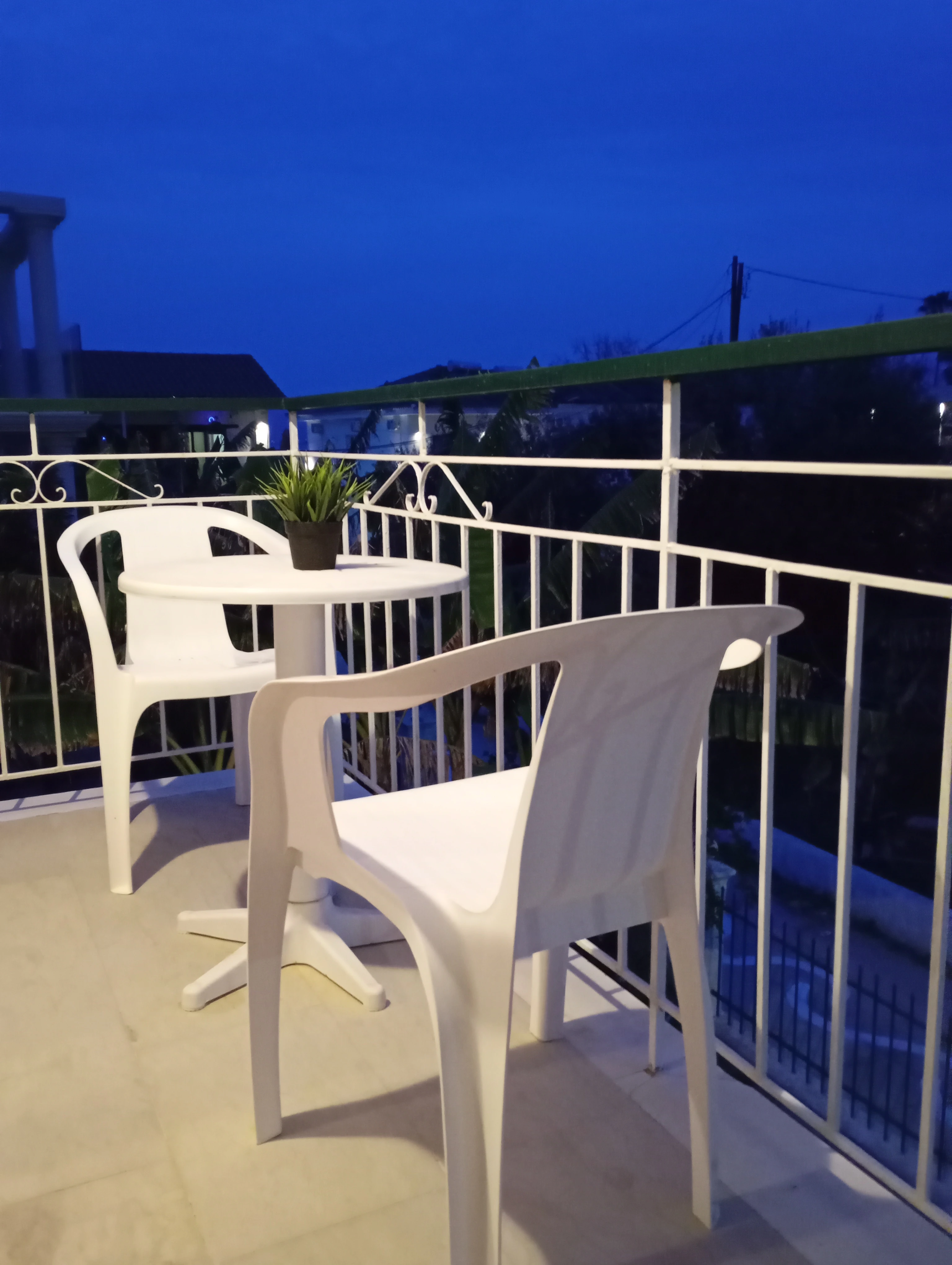 White plastic table and chairs with a potted plant on a balcony overlooking the neighborhood at dusk