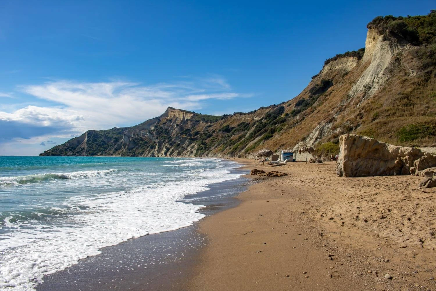 Sandy beach with turquoise waves, dramatic sandstone cliffs covered in green vegetation, and a rustic beach shelter under a